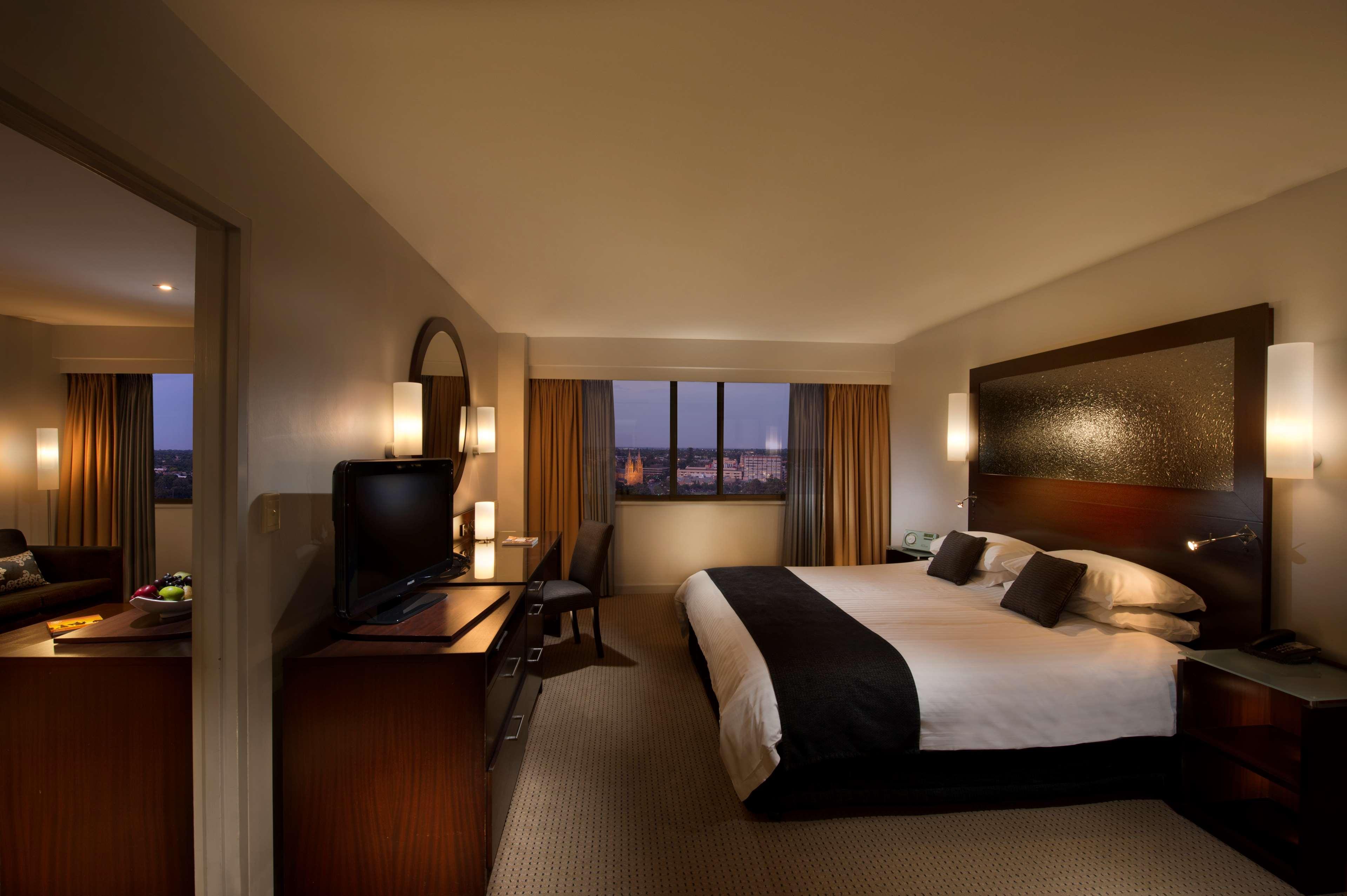 Hotel Stamford Plaza Adelaide Exterior foto The photo features a modern hotel room that includes a large bed with dark and light bedding, complemented by two pillows. The room is well-lit with warm lighting and has a contemporary aesthetic. To one side, there is a wooden dresser with a flat-sc