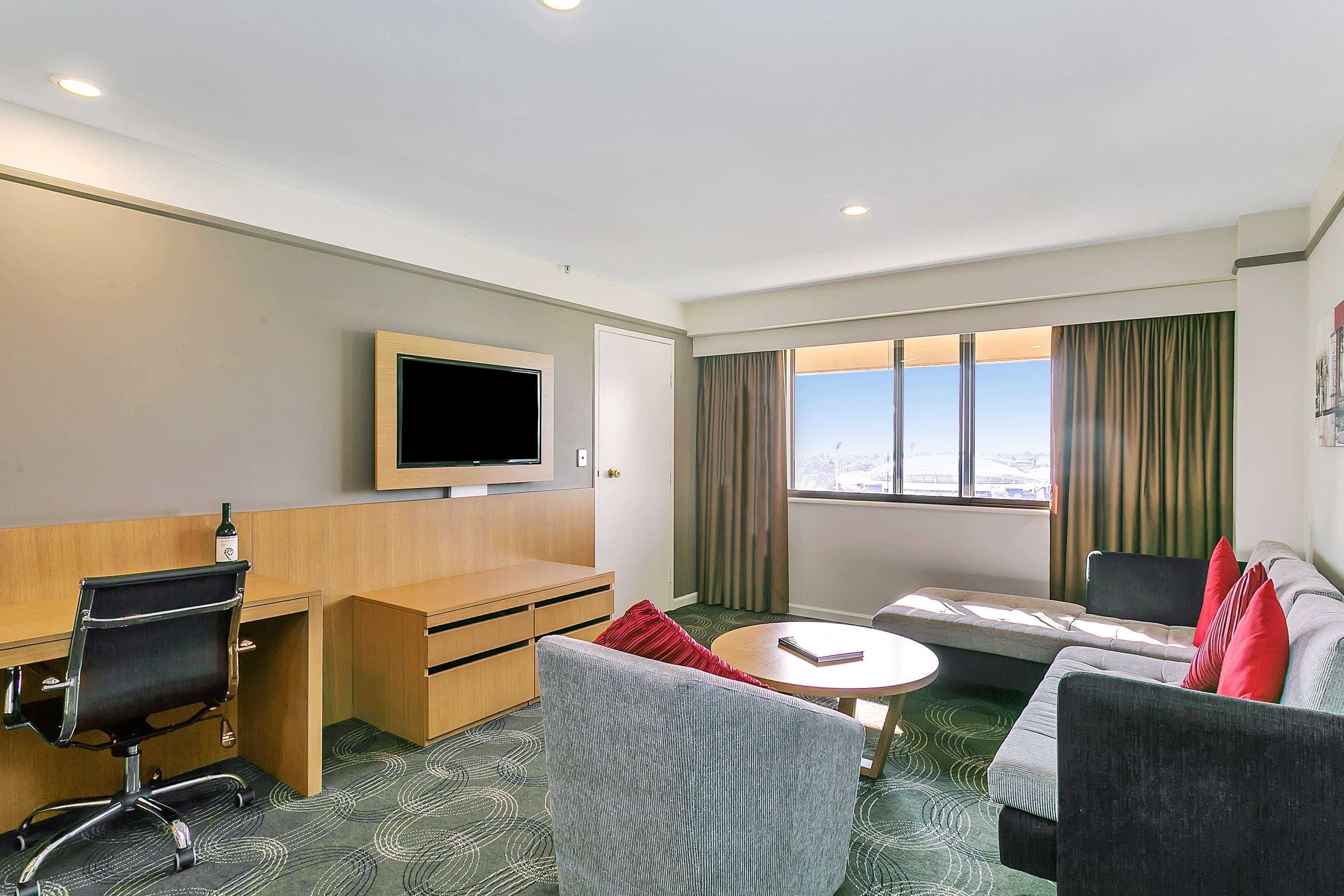 Hotel Stamford Plaza Adelaide Exterior foto The photo shows a modern hotel room or suite that features a cozy seating area. There is a comfortable sofa with red accent pillows, a round coffee table in the center, and a sleek desk with an office chair. On the wall, there is a flat-screen TV mou