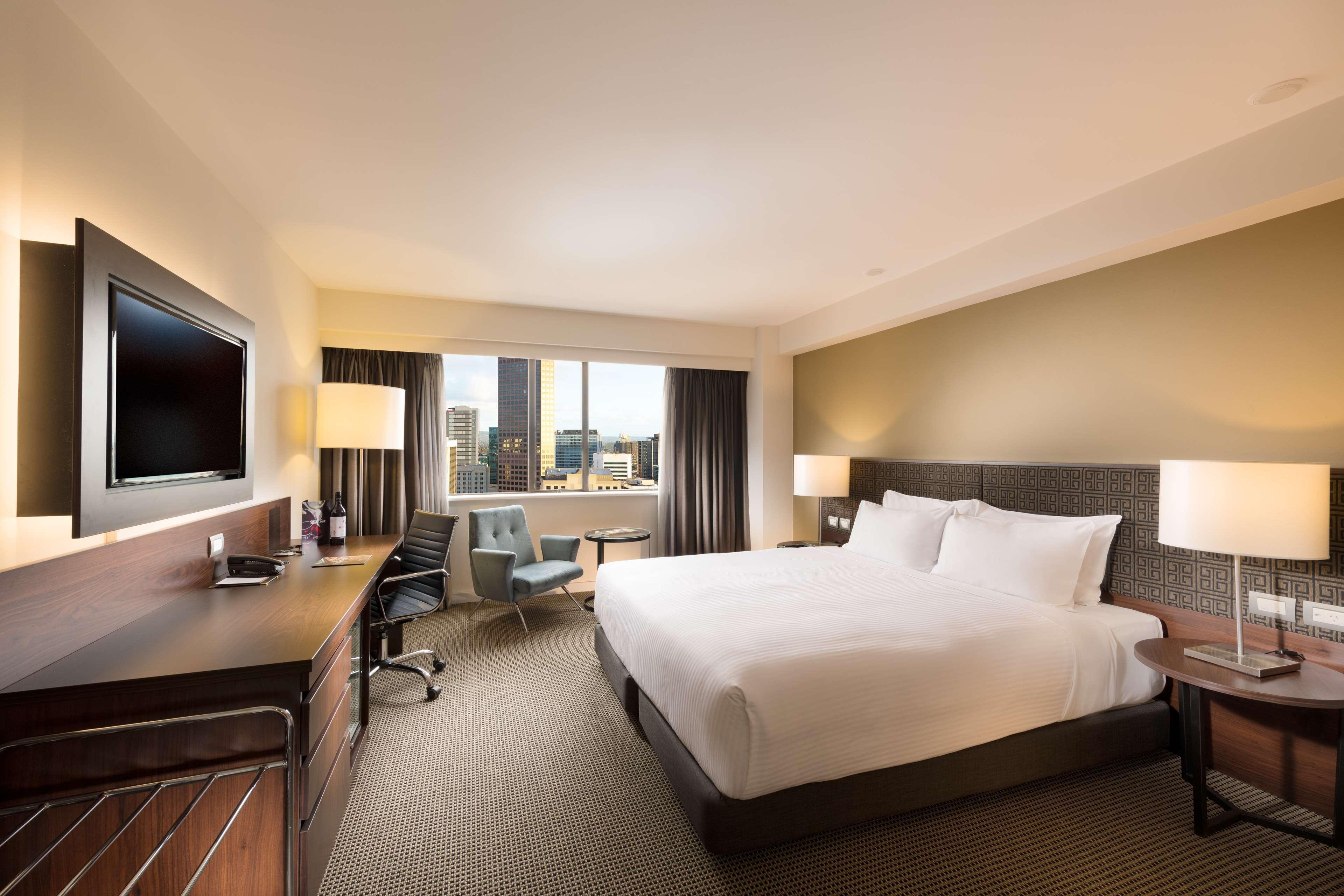 Hotel Stamford Plaza Adelaide Exterior foto The photo shows a modern hotel room with a contemporary design. It features a large bed with white linens, a bedside table on either side with lamps, and a dark wood accent wall. There's a work desk with an office chair and a flat-screen television m